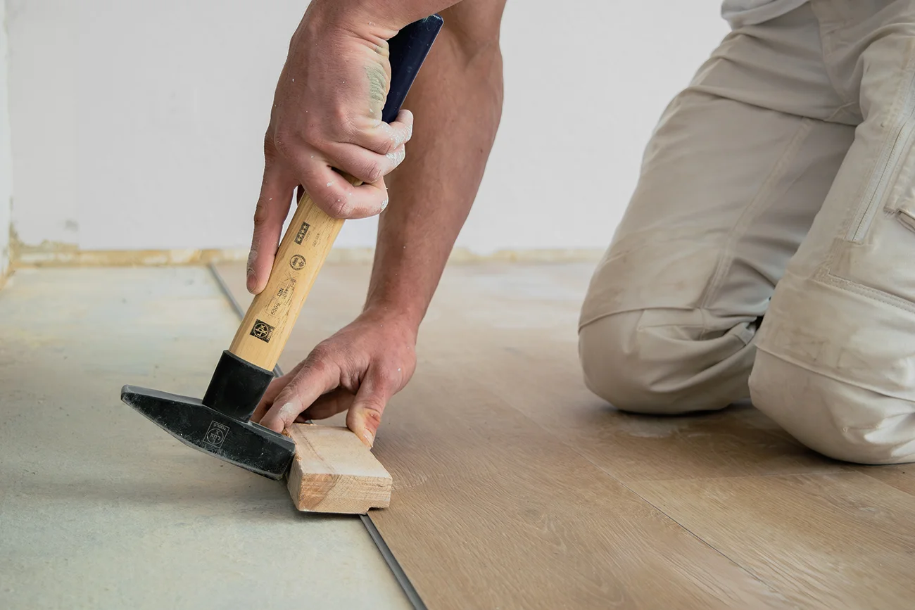 Eine Person kniet auf dem Boden und verlegt mit einem Hammer und einem Holzklotz einen Laminatfußboden. Die Hände des Arbeiters sind zu sehen, wie er die Holzdiele mit idealem Werkzeug an ihren Platz drückt.