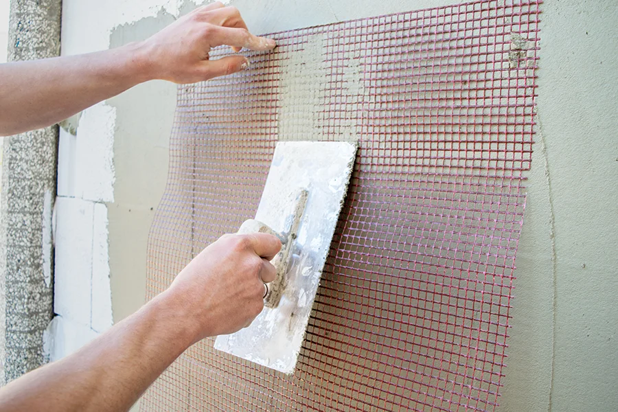 Eine Person glättet mit einer flachen Kelle Putz über ein rotes Glasfasernetz, das an einer Wand befestigt ist. Es wird eine Fassade neu verputzt.