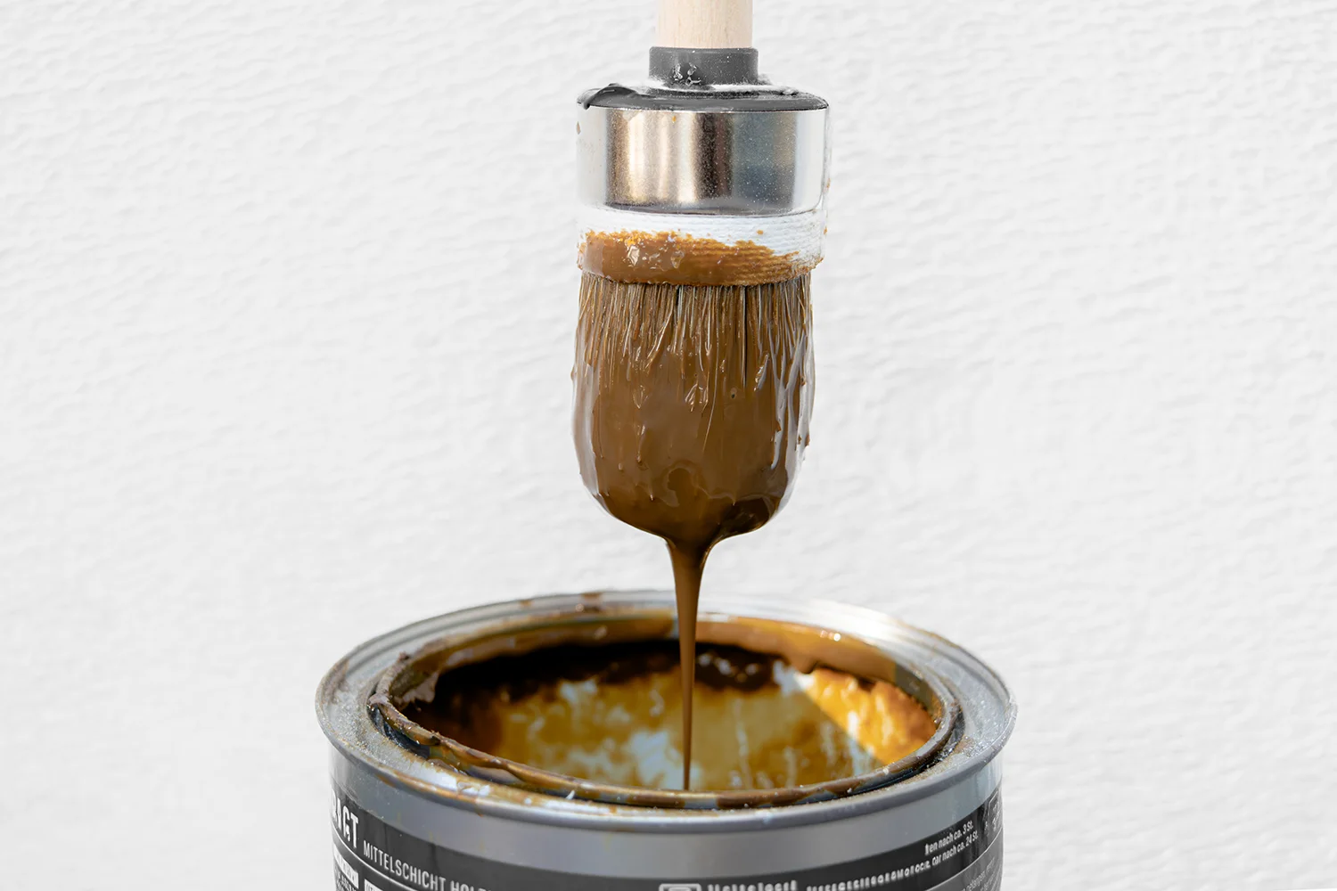Ein mit brauner Lasurfarbe bedeckter Pinsel wird über eine offene Farbdose gehalten, wobei die Farbe zurück in die Dose tropft. Der Hintergrund ist eine einfarbig weiße Fläche.