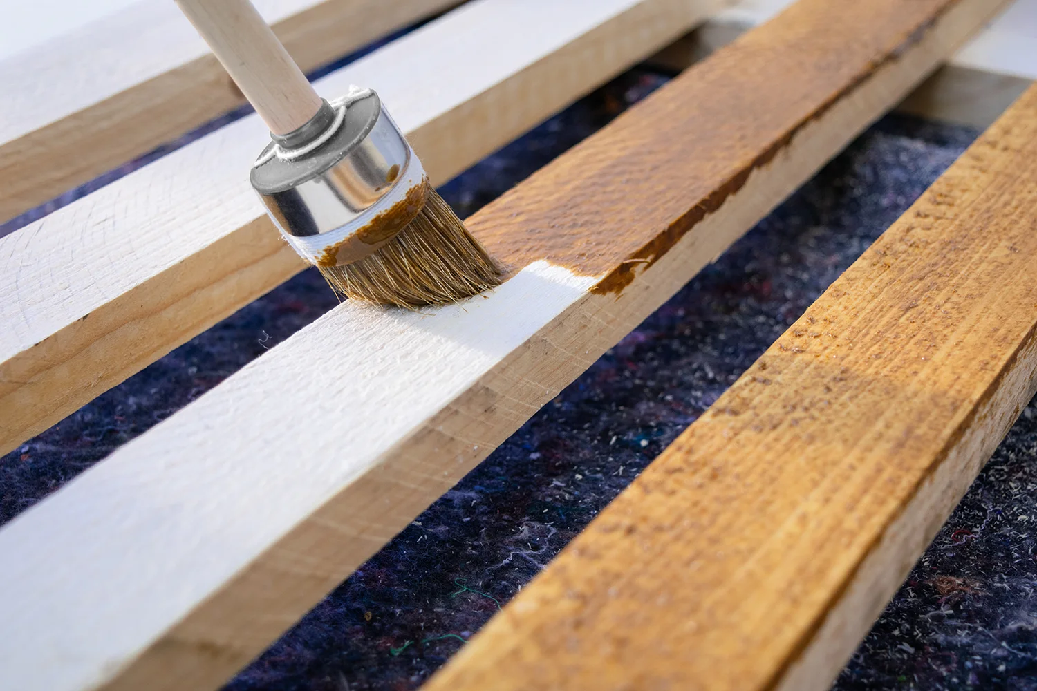 Nahaufnahme eines Pinsels, mit dem braune Lasurfarbe auf eine Holzoberfläche aufgetragen wird. Andere Latten zeigen unbemaltes und teilweise bemaltes Holz. Das Holz wird neu gestrichen, um neuen Schutz zu gewähren.