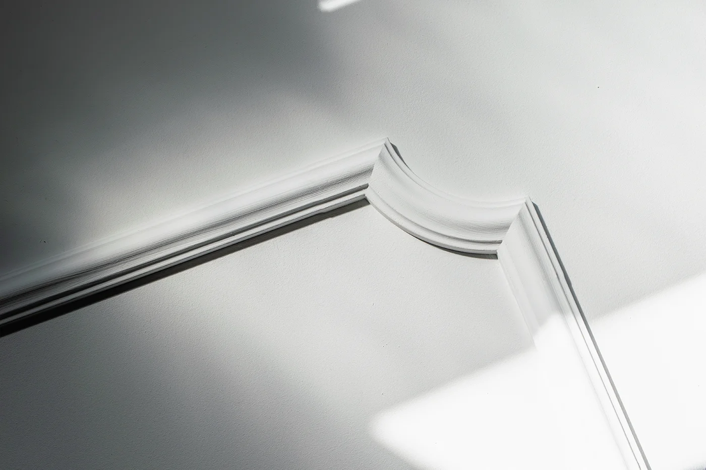 Ein minimalistisches Foto einer weißen Decken mit dekorativen Stuckleisten, beleuchtet durch weiches natürliches Licht und Schatten. Das Bild hebt einfache architektonische Details und geometrische Formen hervor.