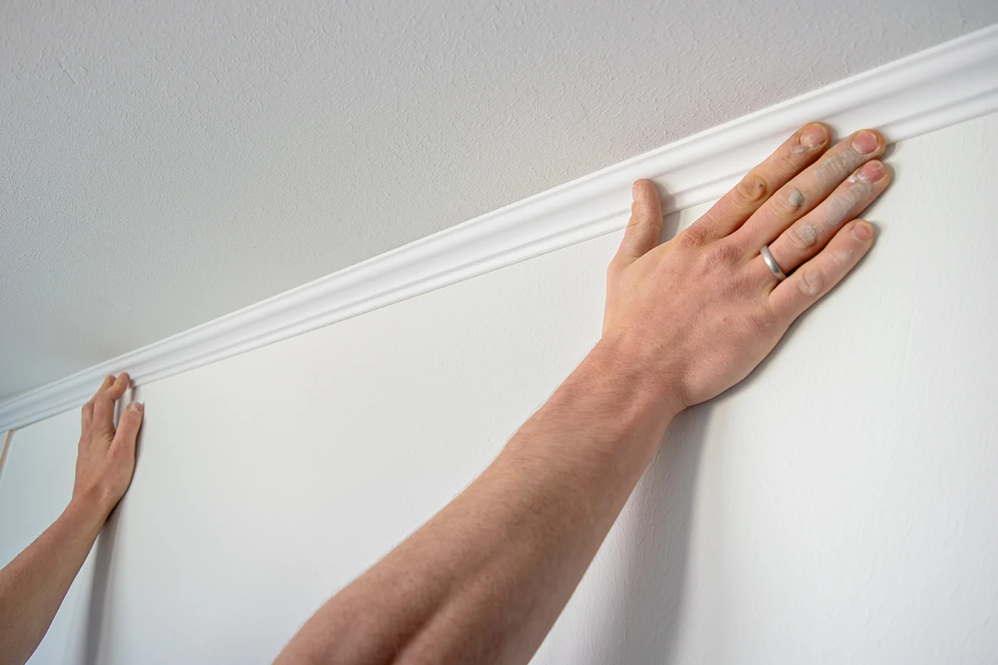 Eine Person bringt eine weiße Zierleiste entlang der Fuge zwischen einer weißen Wand und der Decke an und drückt sie mit beiden Händen fest.