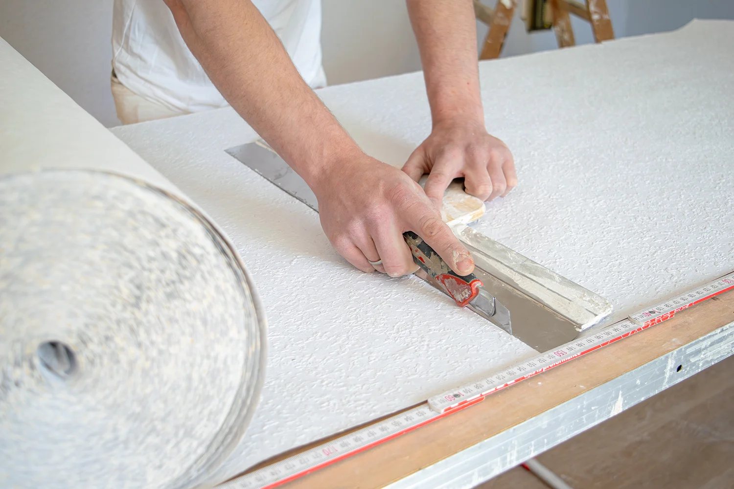 Eine Malermeister aus Bornheim schneidet mit einem Universalmesser und einem Metalllineal Strukturtapete beziehungsweise Raufasertapete auf einem Arbeitstisch zu. Auf dem Tisch ist auch ein Gliedermaßstab zu sehen.