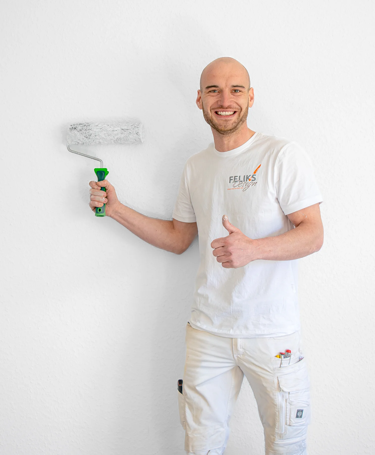 Ein lächelnder Mann in weißer Malerkleidung steht vor einer weißen Wand, hält eine Farbrolle in der einen Hand und zeigt mit der anderen den Daumen nach oben. Die Wand ist weiß und die Farbrolle ist grau. Die Wand wird von dem Mann neu gestaltet. Das ist ein Kundenprojekt im Malerbereich.