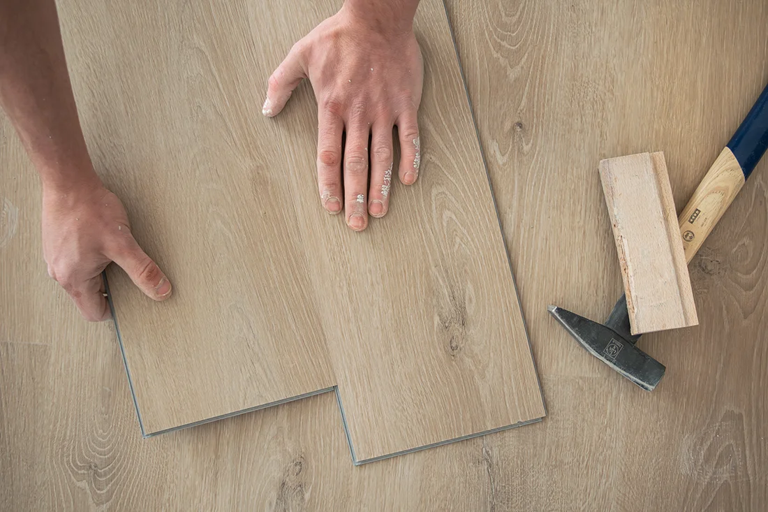 Zwei Hände sind dabei, Vinylboden zu verlegen. Neben den Dielen liegen ein Hammer und ein kleiner Holzklotz auf dem Boden, womit die der Bodenbelag verlegt wird.
